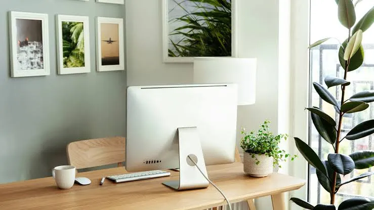 A desk with a gallery wall behind it and a small cream-coloured pot beside the monitor.